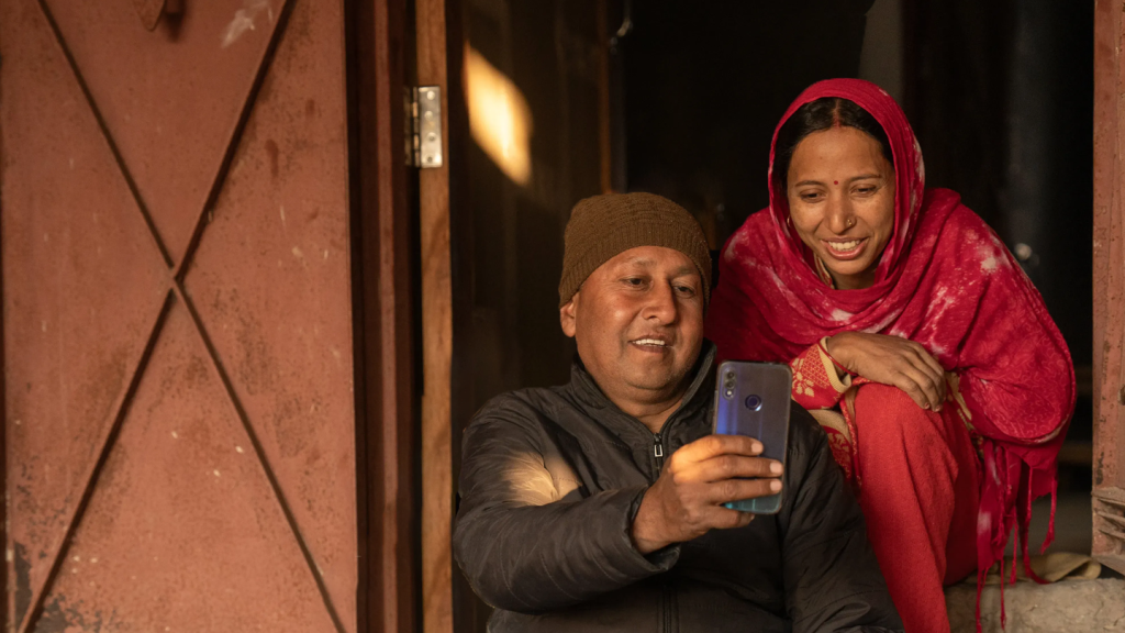 A man in a brown knitted hat and dark jacket sits on a step, smiling as he holds a mobile phone for a selfie. Beside him, a woman in a bright pink sari and scarf crouches, also smiling, with a wooden door and sunlight in the background.
