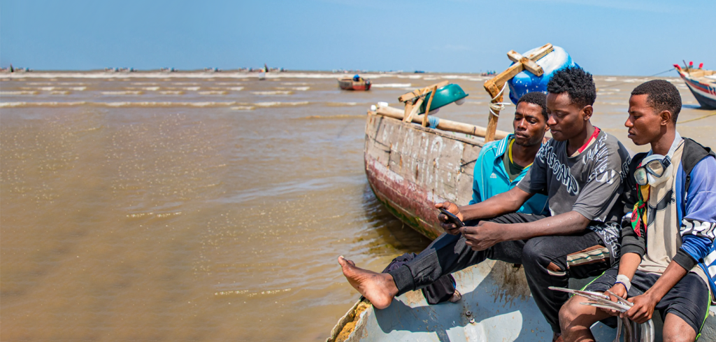 Three young men sit on the edge of a wooden fishing boat near the shore, looking at a device together. The sea is calm with gentle waves and several boats are visible in the background under a clear blue sky.