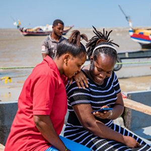 Two women sit outdoors on a jetty, closely looking at a mobile phone and smiling. One wears a red top; the other wears a black-and-white striped dress. A man stands behind them near fishing boats moored on brownish water under a clear blue sky.