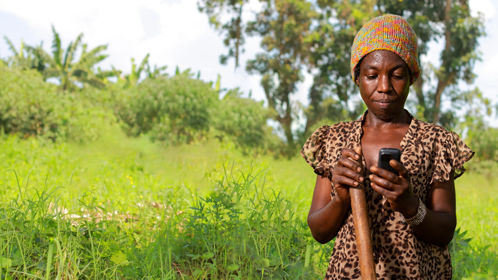 A woman wearing a colourful knitted hat and leopard print shirt stands outdoors in a lush, green field, holding a wooden stick in one hand and using a mobile phone with the other. Trees and dense vegetation are visible in the background under a bright sky.