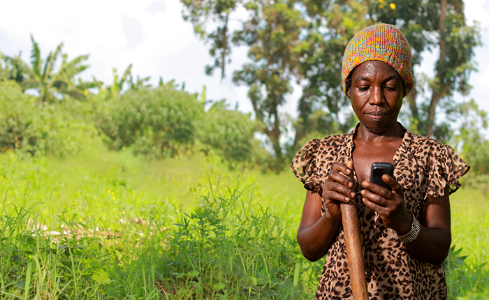 A woman wearing a colourful knitted hat and leopard print top stands outdoors in a lush, green field, looking down at a mobile phone in her hand. She holds a wooden stick and appears focused, with trees and vegetation in the background.
