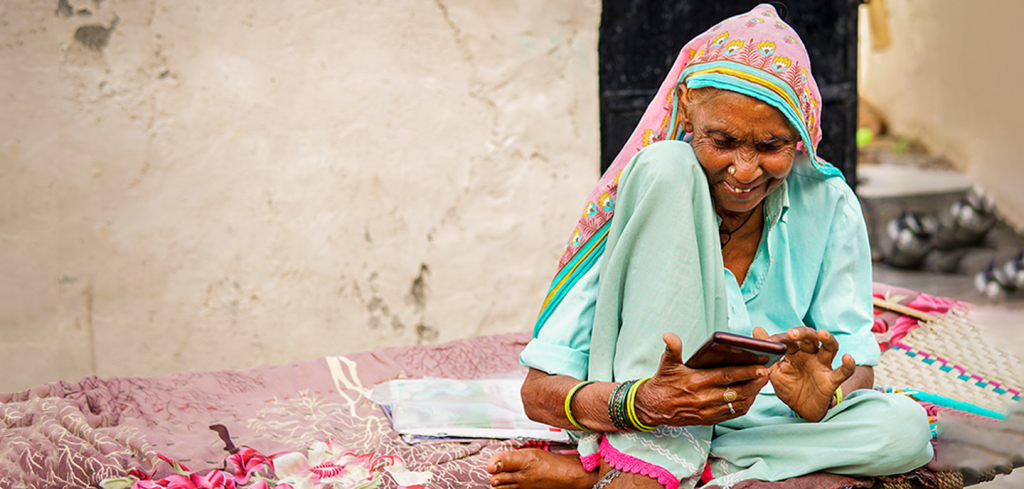 An elderly woman in a colourful headscarf and light blue traditional clothing sits cross-legged on a bed outdoors, smiling as she uses a mobile phone. The background shows a plain wall and some items out of focus. She looks engaged and content.
