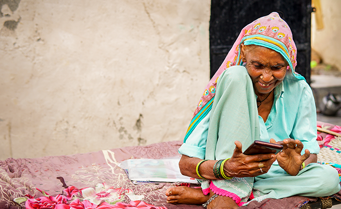 An elderly woman in colourful traditional clothing sits cross-legged on a patterned bed outdoors, smiling as she uses a mobile phone. She wears a light blue outfit, a bright headscarf, and several bangles. The background is a faded wall with a soft, neutral tone.