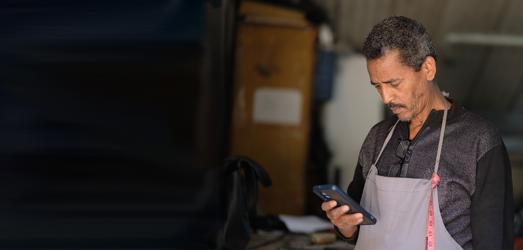A middle-aged man with short curly hair, wearing a dark jumper, grey apron, and pink measuring tape around his neck, stands indoors looking intently at a mobile phone in his hand; background is blurred and partially obscured.