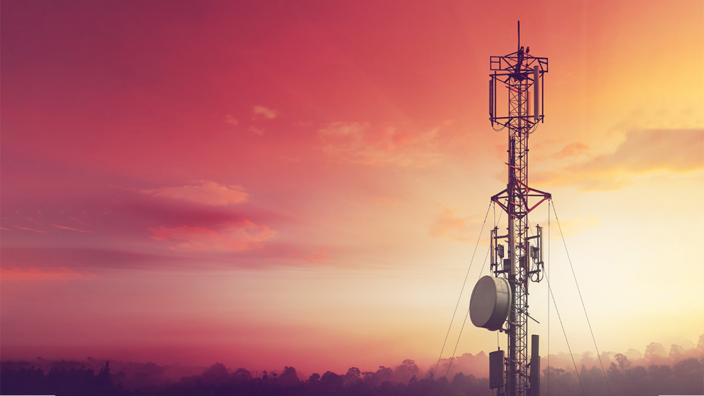A tall mobile mast with multiple aerials and dishes stands against a vibrant sunset sky, tinted with warm shades of orange, red, and pink, with soft clouds and silhouetted treetops on the horizon.