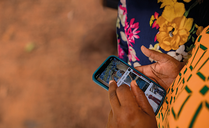 A person wearing a yellow patterned dress uses a mobile phone outdoors. Their hand is touching the screen, browsing photos or social media. Another person in a dark, floral outfit stands nearby. The background is blurry, showing reddish-brown ground.