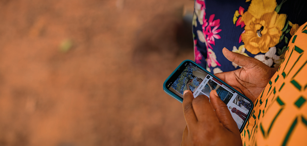 A person wearing patterned clothing uses a mobile phone outdoors. Their hands are visible as they scroll through images on the phone. The background shows blurred reddish-brown ground and part of another person in a brightly coloured floral outfit.