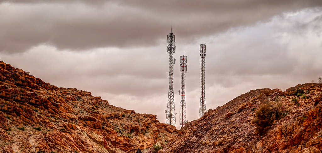 Three tall mobile phone masts stand between rugged, reddish-brown rocky hills under a cloudy sky. The scene is remote and arid, with sparse vegetation on the slopes and dramatic clouds overhead, highlighting the contrast between technology and nature.