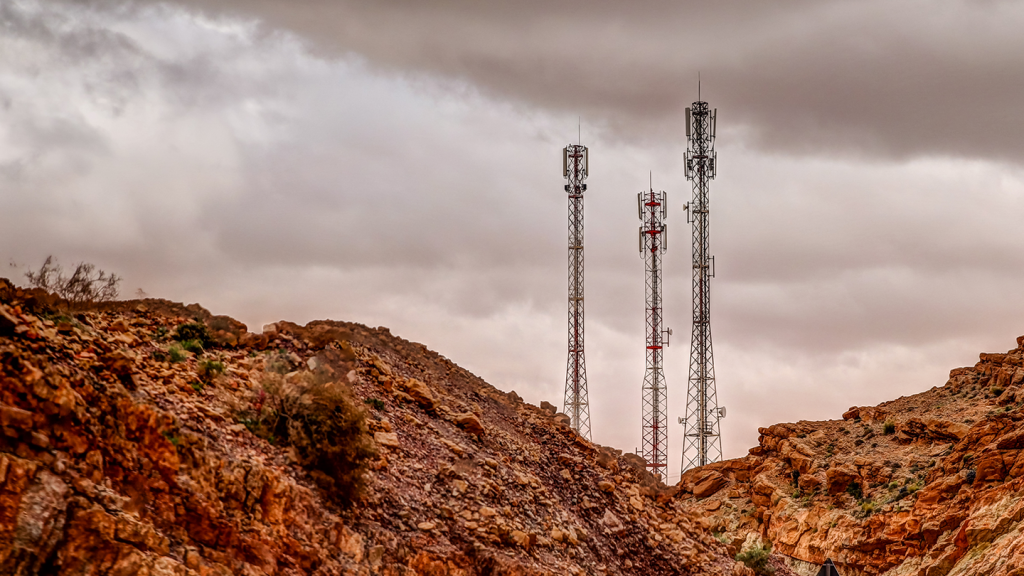 Three tall metal mobile phone masts stand among reddish rocky hills under a cloudy sky. Sparse vegetation dots the rugged landscape, and the masts are topped with aerials and dishes, contrasting sharply with the natural surroundings.