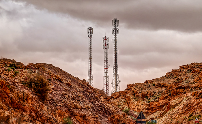 Three tall mobile phone masts stand between rocky, reddish-brown hills under a cloudy, overcast sky. The masts are made of metal and some have aerials or dishes attached. The landscape is rugged, with sparse vegetation on the hills.