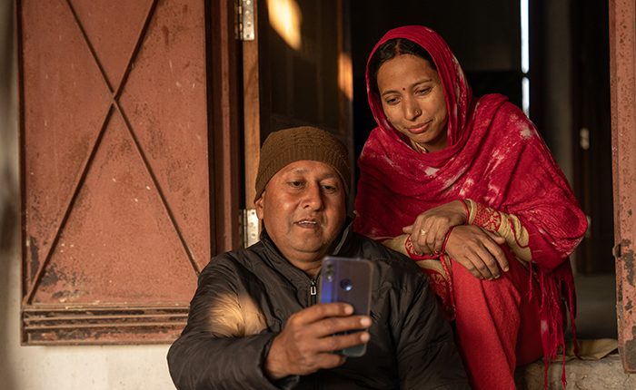 A man in a brown hat and black jacket sits on a doorstep, holding a mobile phone and smiling. Beside him, a woman in a red sari and headscarf looks at the phone, also smiling. They are in warm light, outside a rustic building with a red wooden door.