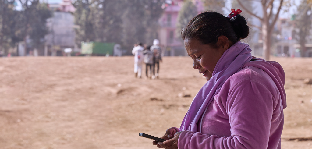 A woman in a pink fleece jacket and scarf stands outdoors, looking at her mobile phone. Her hair is tied back with a red hair clip. The background is a dusty open area with blurred people walking and trees in the distance. The lighting is bright and natural.