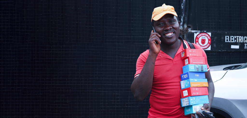 A smiling man in an orange cap and red shirt holds several stacked phone top-up cards and talks on a mobile outside, standing by a white car and a black wall with a red and white sign.
