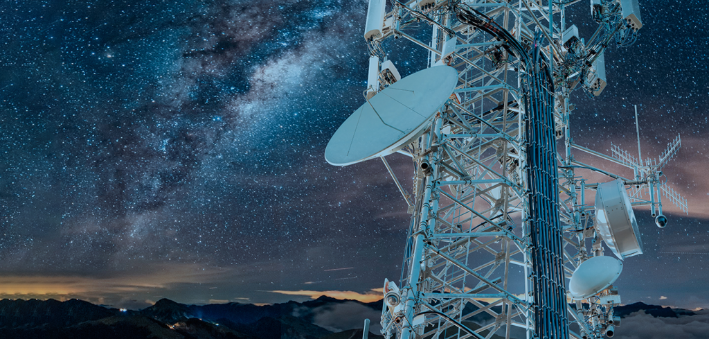 A tall telecommunications tower with multiple aerials and satellite dishes stands against a star-filled night sky, with the Milky Way visible. Mountains and clouds are seen in the distance at the bottom of the image.