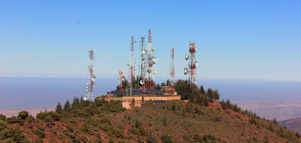 A cluster of telecommunications masts stands atop a mountain, surrounded by trees and green shrubbery. The masts have various aerials and satellite dishes. The sky is clear and blue, and a vast, hazy landscape stretches out in the distance below.
