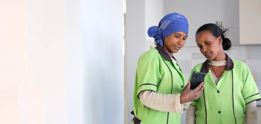 Two women wearing green uniforms stand indoors, closely looking at a mobile phone held by one of them. One woman has a blue headscarf, and both appear focused and engaged with the phone’s screen. The background is bright and minimal.