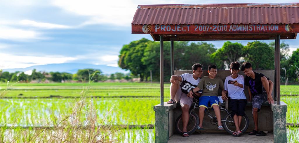 Five young men sit together on a small shelter with a rusty roof, laughing and looking at a mobile phone. Behind them are green rice fields and trees, with mountains in the distance under a blue sky. The shelter sign reads “PROJECT 2007-2010 ADMINISTRATION.”.