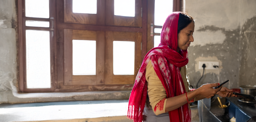 A woman wearing a red and white dupatta stands in a sunlit kitchen near a cooker, holding a cooking utensil. Light streams through a wooden window behind her. The kitchen walls are unpainted and there is a visible power point on the wall.