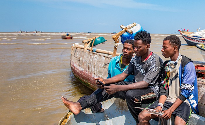 Three young men sit on a weathered wooden boat by the sea, looking at a mobile. They wear casual clothes; one has headphones around his neck. Other boats and people are visible in the background under a clear blue sky.
