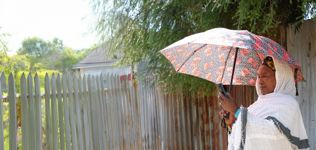 A woman wearing a white traditional garment and headscarf stands outdoors, holding a patterned umbrella for shade. She is near a wooden fence, with trees and sunlight in the background, and looks thoughtfully into the distance.
