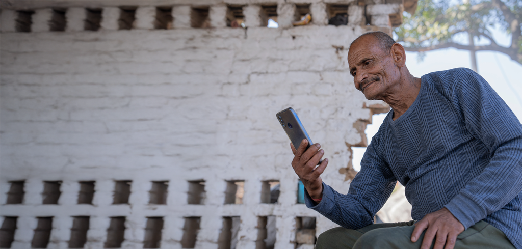 An older man with a moustache, wearing a blue long-sleeved shirt and green trousers, sits outside against a rustic white brick wall. He looks intently at a mobile phone in his hand, with a thoughtful expression. Sunlight and tree branches are visible in the background.