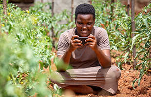 A person with short dark hair, wearing a brown shirt and skirt, sits cross-legged on the ground in a lush garden, surrounded by green plants. They are smiling and looking at a mobile phone held in both hands. The soil is reddish and the day is bright.