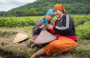 Two women sit smiling in a green field, looking at a mobile phone together. Both wear headscarves and colourful clothing. Traditional conical hats rest beside them. Lush vegetation and trees fill the background under a cloudy sky.