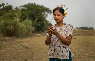 A woman stands outdoors in a grassy field, wearing a patterned shirt and green skirt, with yellow flowers in her hair. She is looking down and holding a mobile phone, with trees and shrubs visible in the background under an overcast sky.