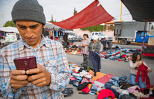 A man in a checked shirt looks at his mobile phone in an outdoor market. Behind him, people sort through piles of clothes spread on the ground under a red canopy. Cars and a lorry are parked along the street in the background.