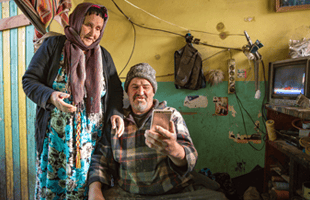 An elderly woman in a headscarf stands beside a seated elderly man in a knitted hat, holding a mobile phone. They are in a small, cluttered, and worn room with green and yellow walls, old electronics, and personal items scattered about.