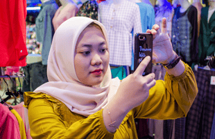 A woman wearing a light hijab and mustard yellow top takes a selfie with her mobile in a clothes shop. She stands in front of rails of colourful clothes and mannequins, focusing intently on capturing her photo.