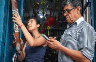 A young woman examines a tall, decorated wooden item in a shop, while an older man in glasses and a striped shirt looks at a mobile. The shop is filled with various objects and decorations, including red lanterns and plants, creating a busy, eclectic atmosphere.
