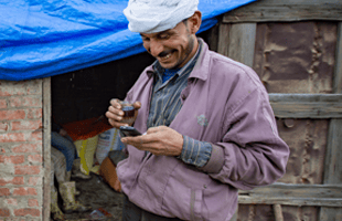 A man in a light purple jacket and white headscarf smiles whilst looking at a phone in his hand. He stands outside a rustic brick building with a worn wooden door and a blue tarpaulin roof. Another person is partially visible sitting inside.