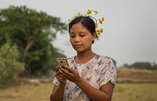 A young woman with brown hair and a yellow flower crown stands outdoors, wearing a light floral dress. She looks down, focused on her mobile phone, with trees and a grassy field blurred in the background. The lighting is soft and natural.
