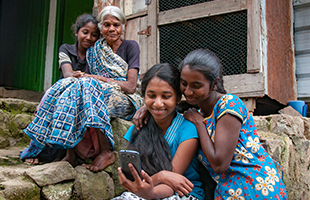 Three women and a young girl sit on stone steps outdoors, smiling and gathered closely as the girl in front holds up a mobile for a selfie. They wear colourful, patterned clothing with a rustic wooden building in the background.