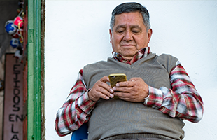 An older man with short grey hair and a serious expression sits and looks at a smartphone. He wears a red checked shirt and beige jumper vest. He is seated by a green door, with a white wall behind him and a wooden sign partially visible on the left.
