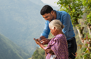 A smiling man in a blue shirt helps an elderly woman, dressed in a purple patterned sari, use a mobile phone outdoors. They stand on a hillside with lush green trees and misty mountains visible in the background.