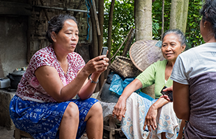 Three women sit outdoors surrounded by trees and baskets. Two women, one in a red patterned shirt and one in a green shirt, are looking at mobile phones. The mood is relaxed and they appear to be sharing something on their devices.