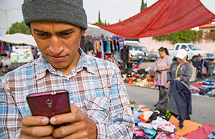 A man in a grey beanie and checked shirt looks intently at his mobile at an outdoor market. Behind him, people browse clothes and items laid out on the ground, with red tarpaulins and vehicles in the background.