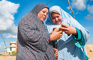 Two women in patterned hijabs stand outdoors under a blue sky with clouds, smiling as they look at a mobile phone together. One wears a grey hijab, the other light blue. Sandy ground and buildings are visible in the background.