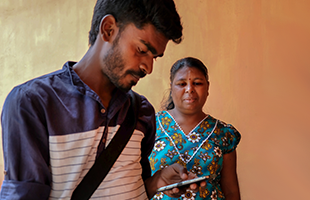 A man with a beard looks down and uses a mobile phone, wearing a navy and white shirt with a bag strap across his chest. Behind him, a woman in a blue floral dress stands against a peach-coloured wall, looking in his direction with a neutral expression.