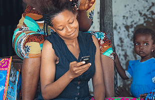 A woman smiles while looking at a mobile as another person styles her hair. A young child in a blue outfit stands nearby, watching. Bright coloured clothing and a peeling wall are visible in the background. The mood appears relaxed and informal.