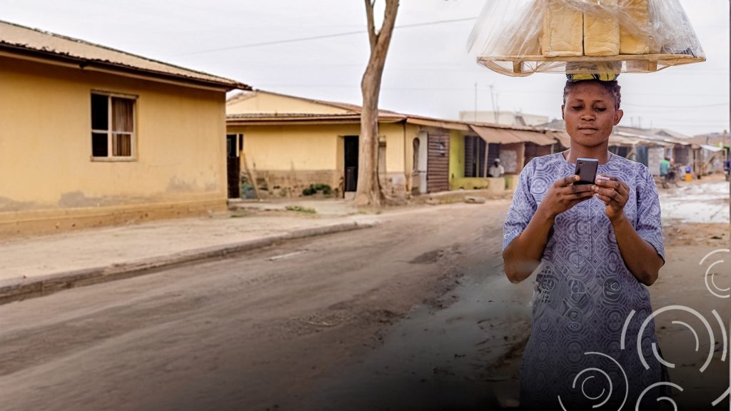 A woman in a patterned dress balances a tray of bread on her head while using a mobile phone, walking along a quiet, dusty street lined with yellow houses. The background shows overcast skies and an empty road in a residential neighbourhood.