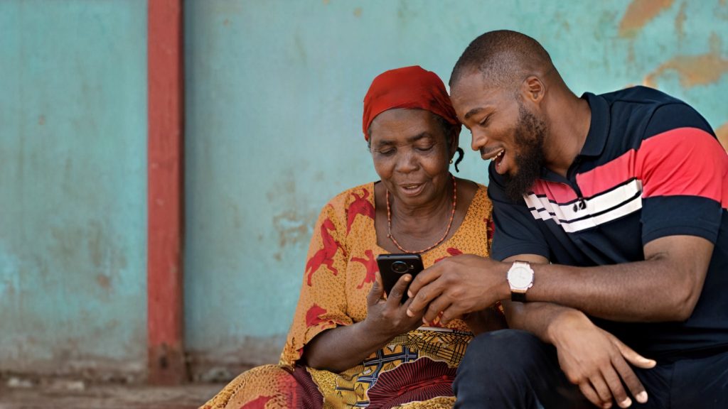An elderly woman in a patterned dress and red headscarf sits beside a younger man in a striped polo shirt. They smile and look at a mobile phone the woman is holding, sitting against a blue wall with a red pillar.