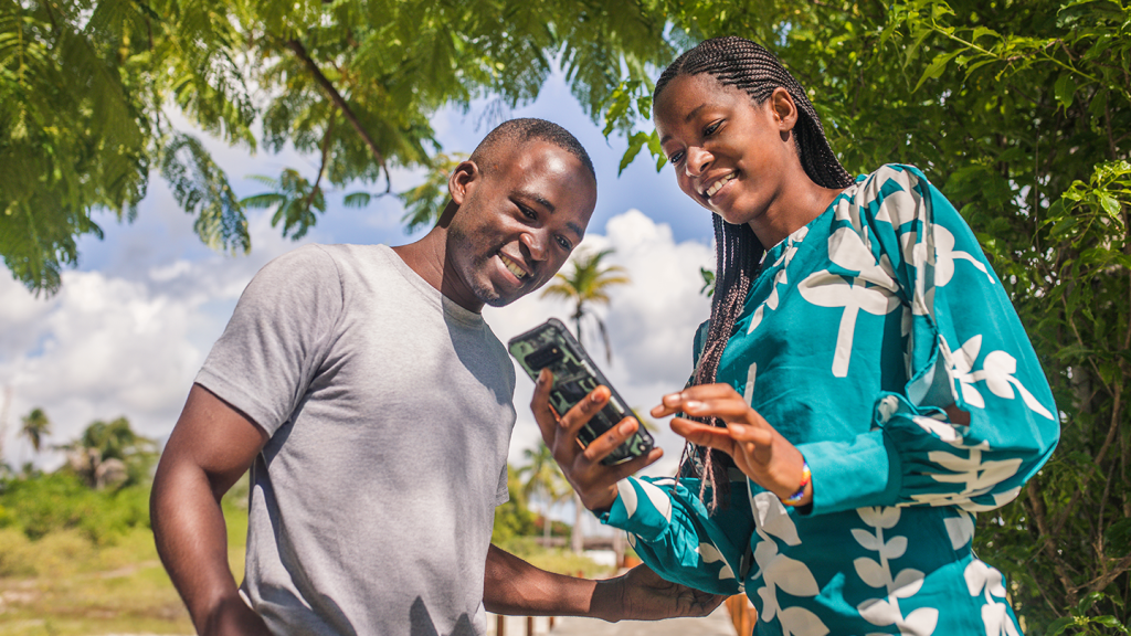 A smiling man in a grey T-shirt and a woman in a blue patterned blouse look at a smartphone together outdoors. Green trees, palm trees, and a bright sky are visible in the background.