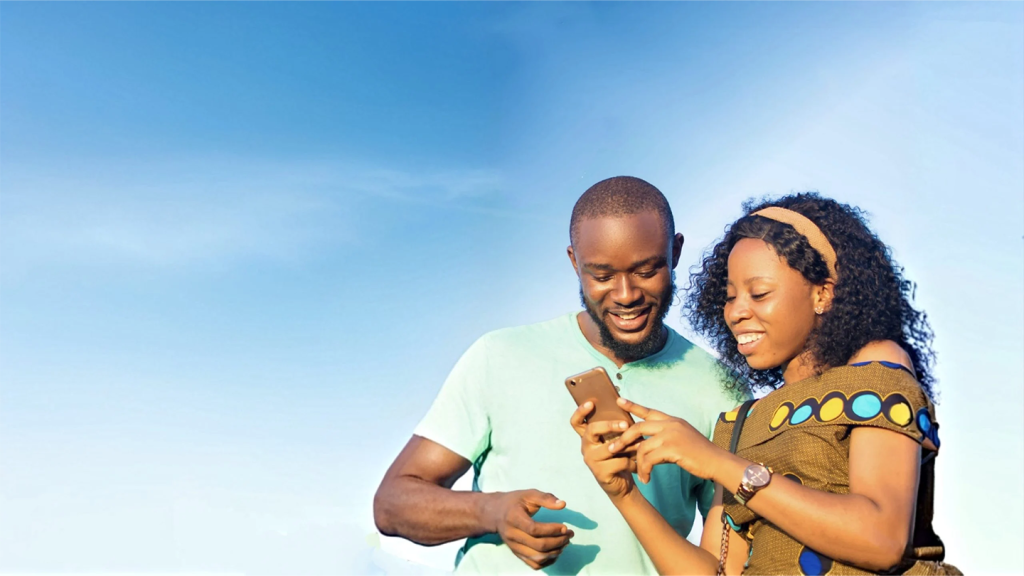 A smiling man and woman stand together outdoors against a bright blue sky. The woman holds a mobile phone, and both appear engaged and happy as they look at the phone screen. The scene feels warm and joyful, suggesting a positive moment.