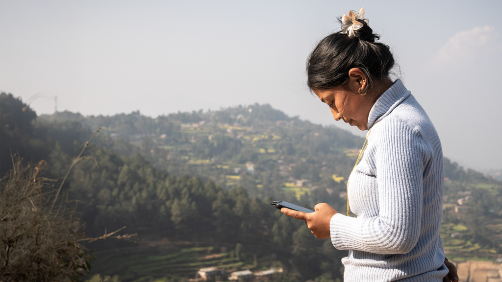 A woman with dark hair tied in a bun looks down at a mobile phone she is holding against a backdrop of hazy, terraced green mountains. She wears a light-blue, ribbed polo-neck jumper and a small hair accessory.