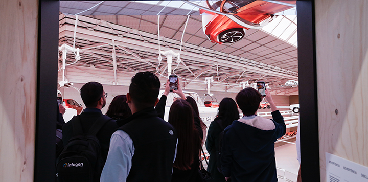 People watching and photographing a display of upside-down cars and a goat in an exhibition hall.