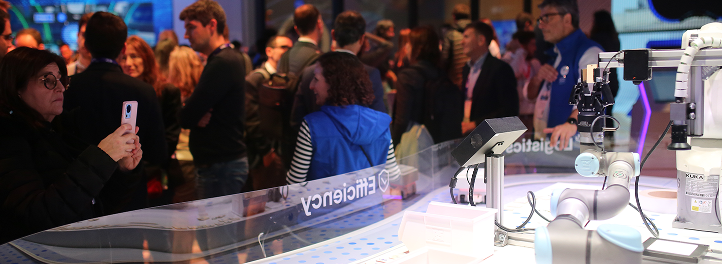 Crowd observes robotics demonstration behind clear barrier at a technology expo. a woman in the forefront takes a photo with her phone.