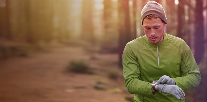 A person wearing a green long-sleeve shirt, a gray beanie, and gloves is looking at a watch on their wrist in a forested area. The surrounding environment appears blurred, with tall trees and a lit path visible in the background.